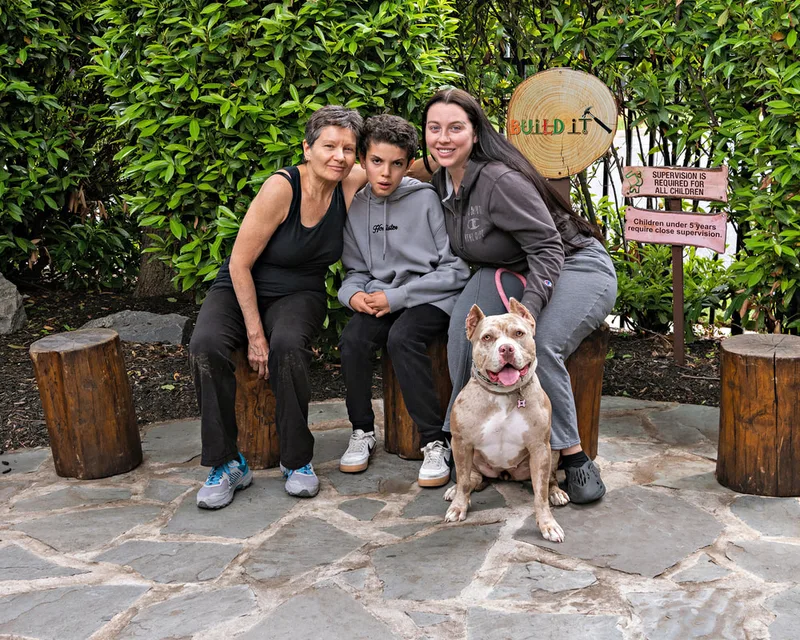 A color photo of a pit bull dog and his family. The photo was taken by pet photographer Laura Paesano of Laura Pea Photography for a pet portrait program held by the Middle Country Public Library in their outdoor Nature Explorium. 