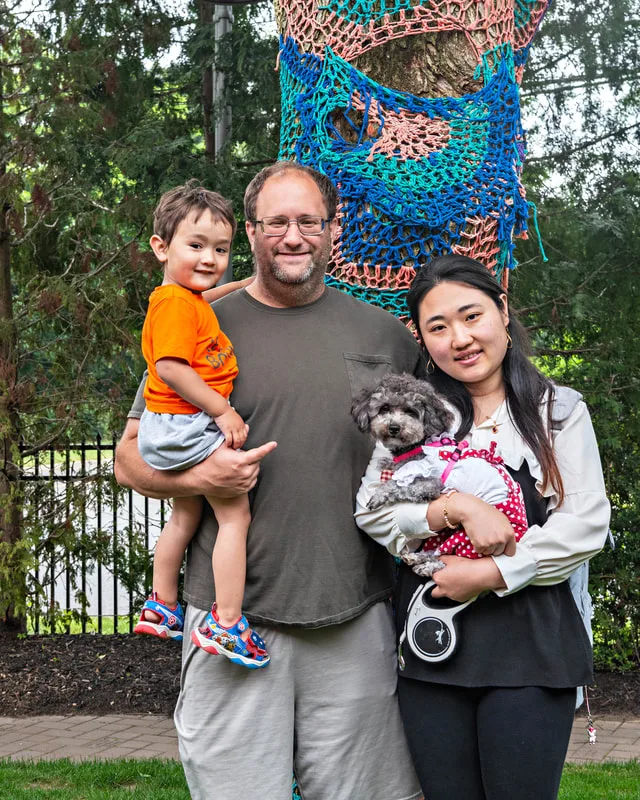 A color photo of a poodle dog and family. The photo was taken by pet photographer Laura Paesano of Laura Pea Photography for a pet portrait program held by the Middle Country Public Library in their outdoor Nature Explorium. 