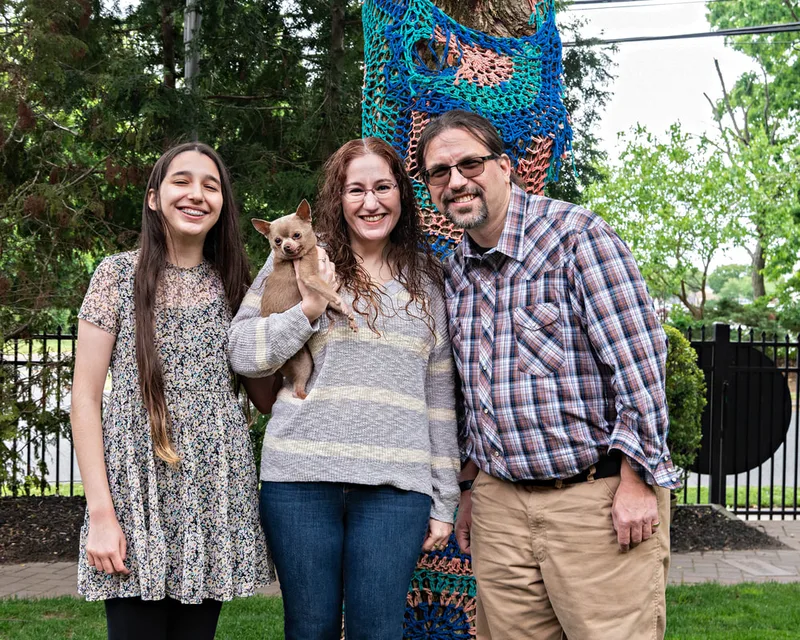 A color photo of a chihuahua dog with its family. The photo was taken by pet photographer Laura Paesano of Laura Pea Photography for a pet portrait program held by the Middle Country Public Library in their outdoor Nature Explorium. 
