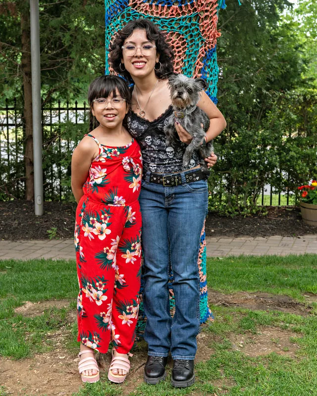 A color photo of a terrier mix dog and two girls. The photo was taken by pet photographer Laura Paesano of Laura Pea Photography for a pet portrait program held by the Middle Country Public Library in their outdoor Nature Explorium. 