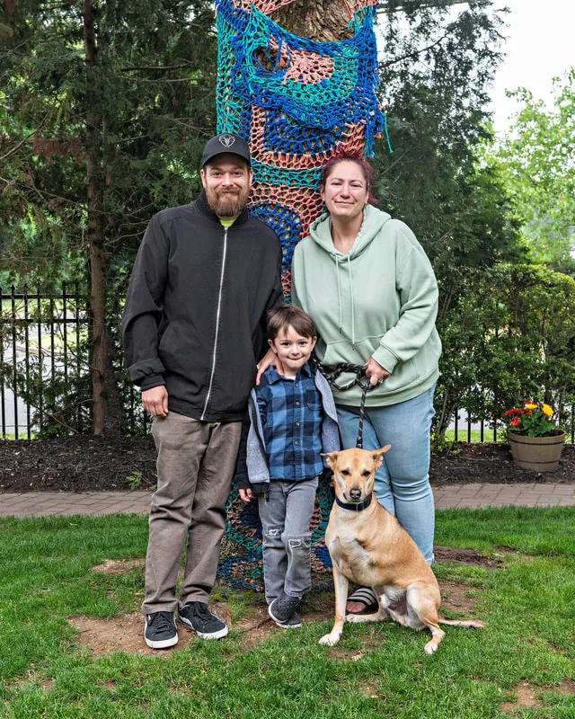 A color photo of a Thai ridgeback dog with three legs with a man, woman and a little boy. The photo was taken by pet photographer Laura Paesano of Laura Pea Photography for a pet portrait program held by the Middle Country Public Library in their outdoor Nature Explorium. 