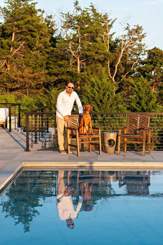 color photo of a red vizsla dog in her owner's backyard on the water. Her and her owner are posing behind a pool