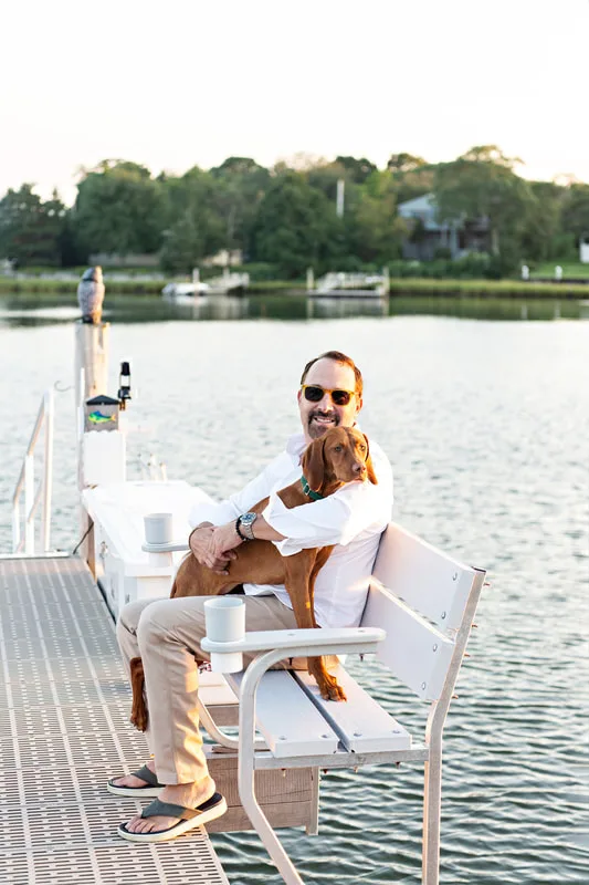 color photo of a red vizsla dog in her owner's backyard on the water. They're sitting on a bench on a dock