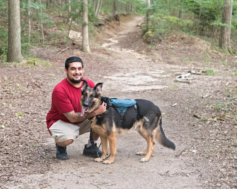 Color photo of a German Shepherd Dog in the woods with his owner. The photo was taken by Laura Paesano of Laura Pea Photography, a Long Island based pet photographer. 