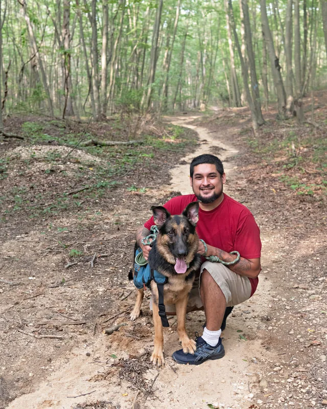 Color photo of a German Shepherd Dog in the woods with his owner. The photo was taken by Laura Paesano of Laura Pea Photography, a Long Island based pet photographer. 