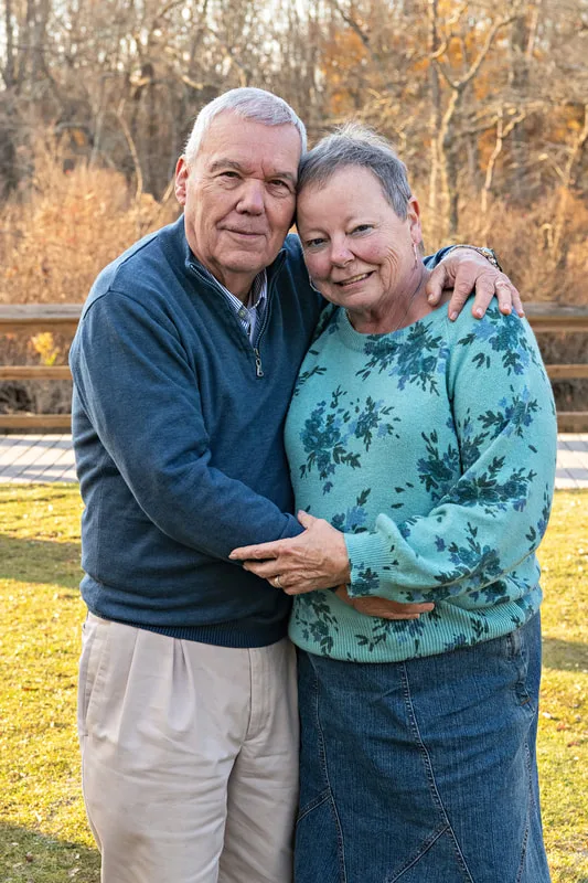 Color photo of a husband and wife hugging each other standing outside on a lawn. 