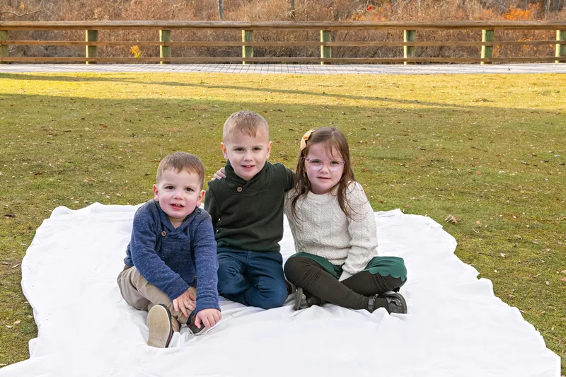 Color photo of a three kids sitting on a white blanket on the grass. 