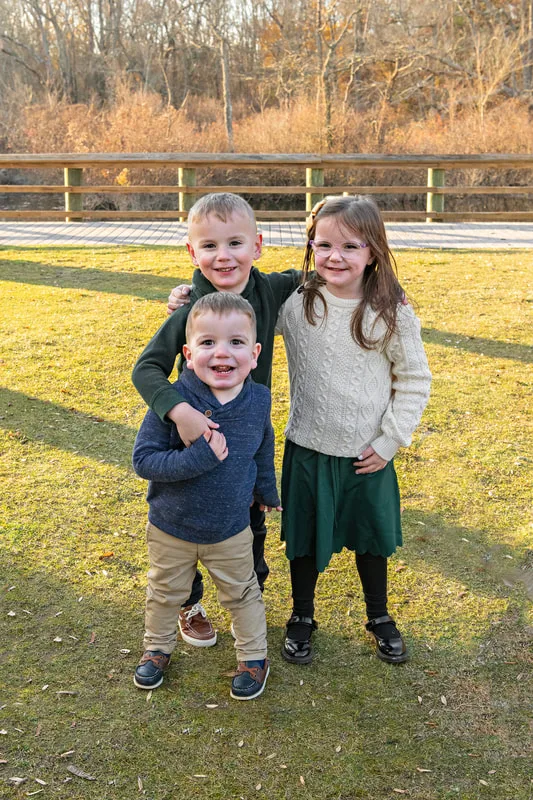 Color photo of a twin boy and girl and their younger toddler brother standing outside on a lawn.