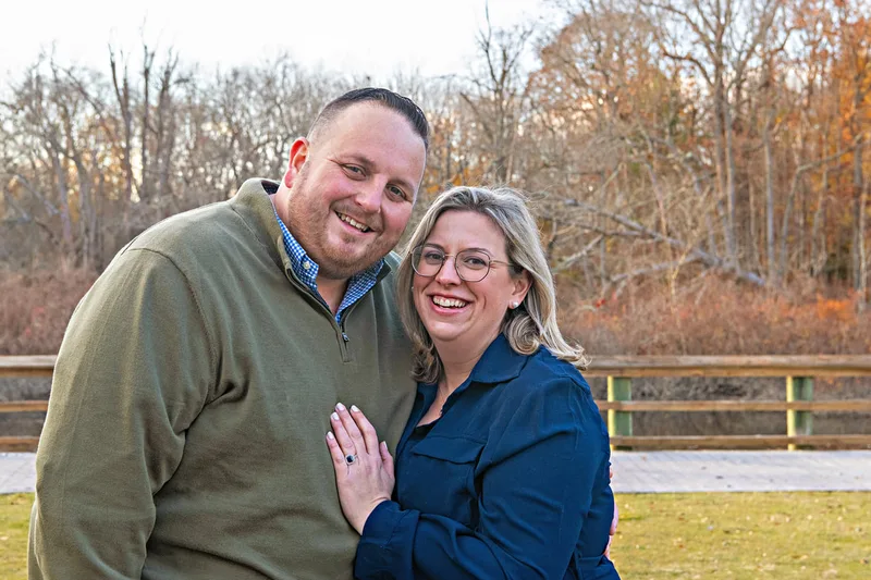 Color photo of a husband and wife posing together standing outside on a lawn. 