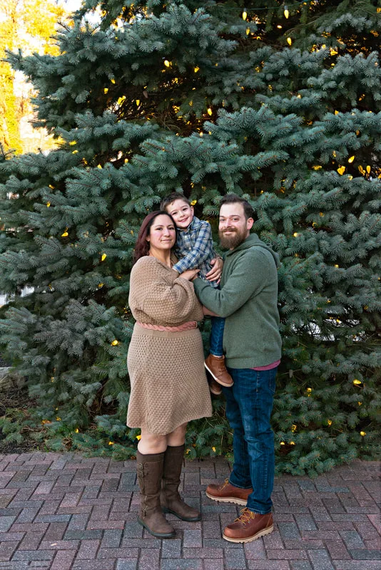 A color photo of a white family of three, a mom, dad and four year old boy in front of a lit Christmas tree. The photo is by Laura Pea Photography, a Long Island based photographer. 
