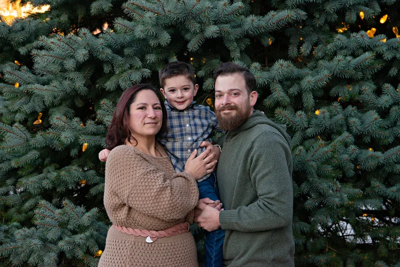 A color photo of a white family of three, a mom, dad and four year old boy in front of a lit Christmas tree. The photo is by Laura Pea Photography, a Long Island based photographer. 