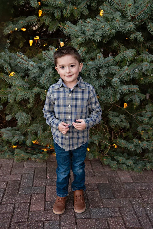 A color photo of a white four year old boy in front of a lit Christmas tree. The photo is by Laura Pea Photography, a Long Island based photographer. 