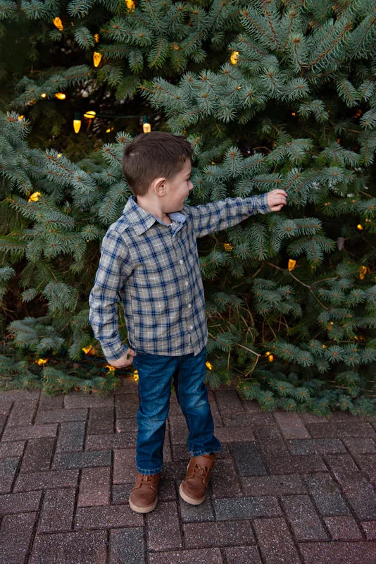 A color photo of a white four year old boy in front of a lit Christmas tree. The photo is by Laura Pea Photography, a Long Island based photographer. 