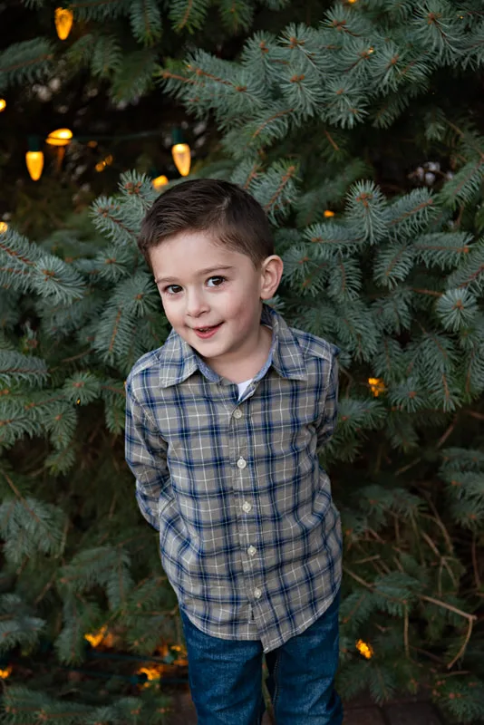 A color photo of a white four year old boy in front of a lit Christmas tree. The photo is by Laura Pea Photography, a Long Island based photographer. 