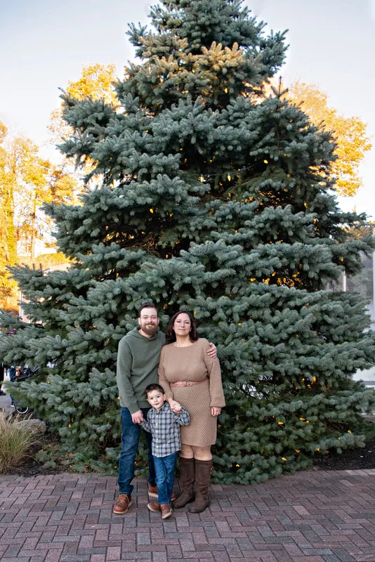 A color photo of a white family of three, a mom, dad and four year old boy in front of a lit Christmas tree. The photo is by Laura Pea Photography, a Long Island based photographer. 
