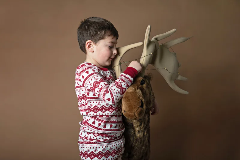 A color photo of a four year old white boy looking at a big stuffed moose in front of a light brown paper backdrop. The photo was taken by Laura Pea Photography, a Long Island based photographer. 