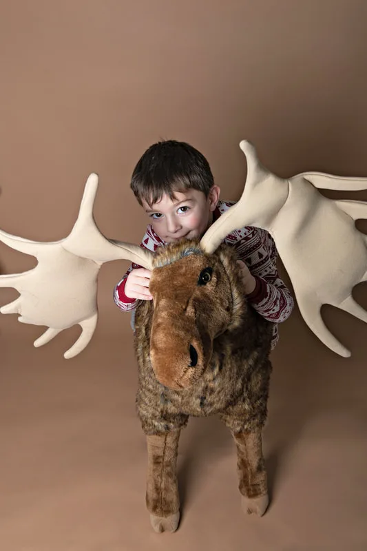 A color photo of a four year old white boy sitting on top of a big stuffed moose in front of a light brown paper backdrop. The photo was taken by Laura Pea Photography, a Long Island based photographer. 