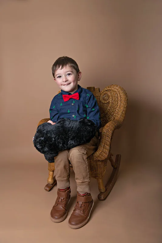 A color photo of a four year old white boy sitting in a wicker chair holding a stuffed bear in front of a light brown paper backdrop. The photo was taken by Laura Pea Photography, a Long Island based photographer. 
