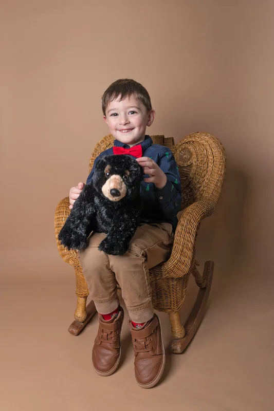 A color photo of a four year old white boy sitting in a wicker chair holding a stuffed bear in front of a light brown paper backdrop. The photo was taken by Laura Pea Photography, a Long Island based photographer. 