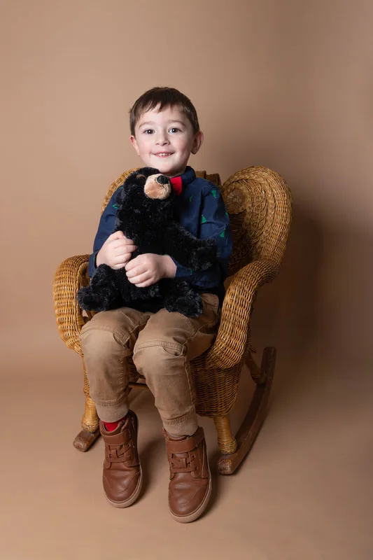 A color photo of a four year old white boy sitting in a wicker chair holding a stuffed bear in front of a light brown paper backdrop. The photo was taken by Laura Pea Photography, a Long Island based photographer. 