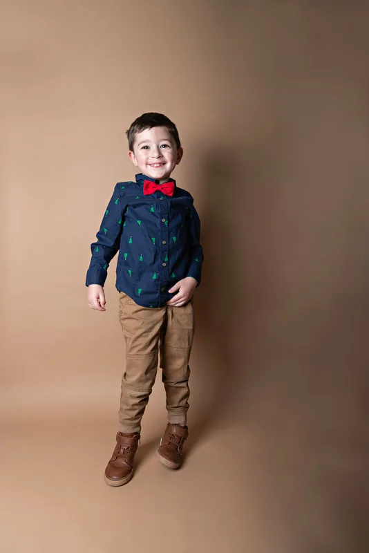 A color photo of a four year old white boy standing in front of a light brown paper backdrop. The photo was taken by Laura Pea Photography, a Long Island based photographer. 