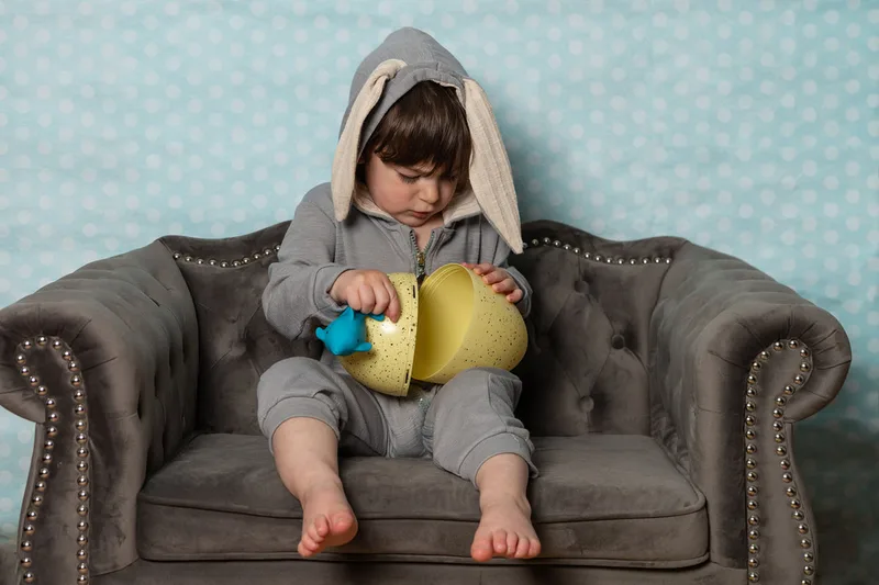 Toddler boy dressed in an Easter bunny costume holding a big yellow egg cracking it in half and looking down into it
