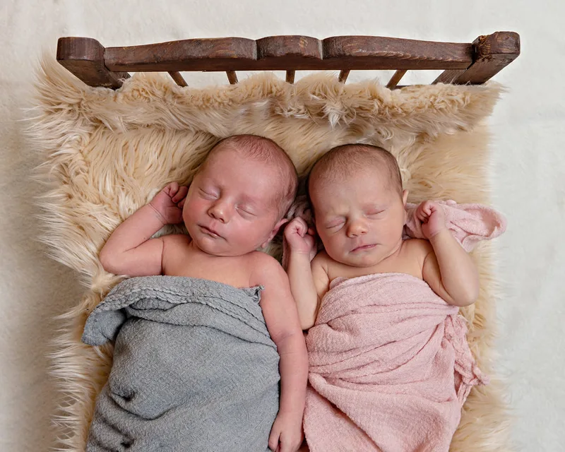 photo of a set of newborn baby twins, one boy and one girl. They are wrapped in grey and pink wraps, laying on a beige furry rug on a vintage wooden doll bed