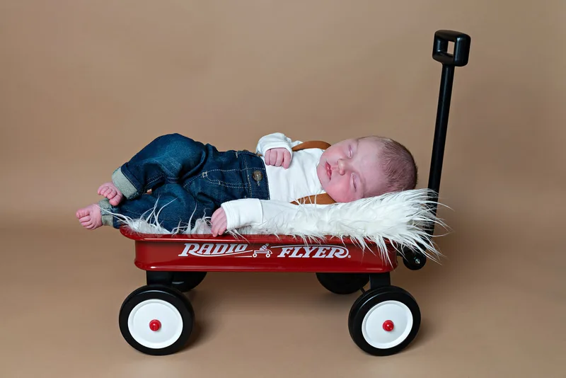 photo of a newborn baby boy in overalls and denim jeans laying on a furry white rug in a red Radio Flyer wagon against a beige paper backdrop in a photo studio