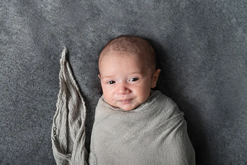 Color close up photo of a newborn baby boy wrapped in a gray wrap on top of a gray blanket photographed in a studio. His eyes are open. The photo was taken by Laura Pea Photography, a Long Island based newborn photographer.