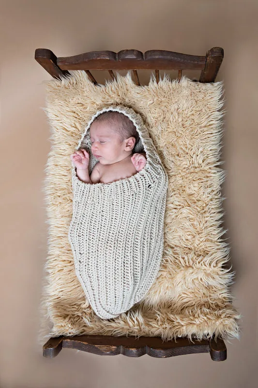 Color photo of a newborn baby boy covered in a beige cocoon wrap on top of a beige furry rug in a vintage doll bed photographed in a studio. His eyes are closed. The photo was taken by Laura Pea Photography, a Long Island based newborn photographer.