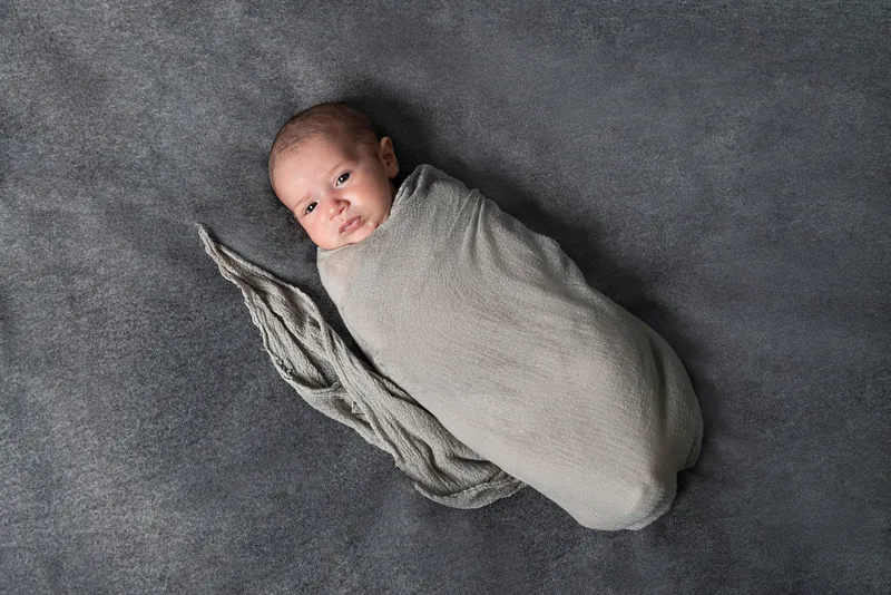 Color photo of a newborn baby boy wrapped in a gray wrap on top of a gray blanket photographed in a studio. His eyes are open. The photo was taken by Laura Pea Photography, a Long Island based newborn photographer.