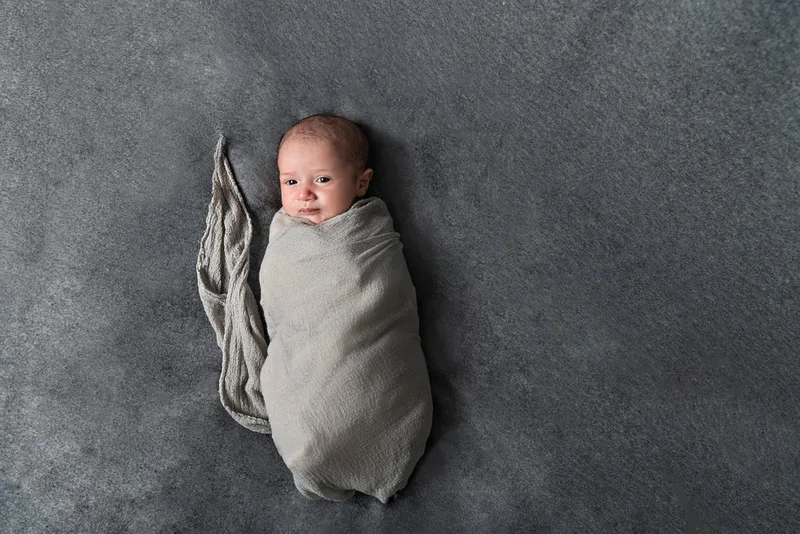 Color photo of a newborn baby boy wrapped in a gray wrap on top of a gray blanket photographed in a studio. His eyes are open. The photo was taken by Laura Pea Photography, a Long Island based newborn photographer.
