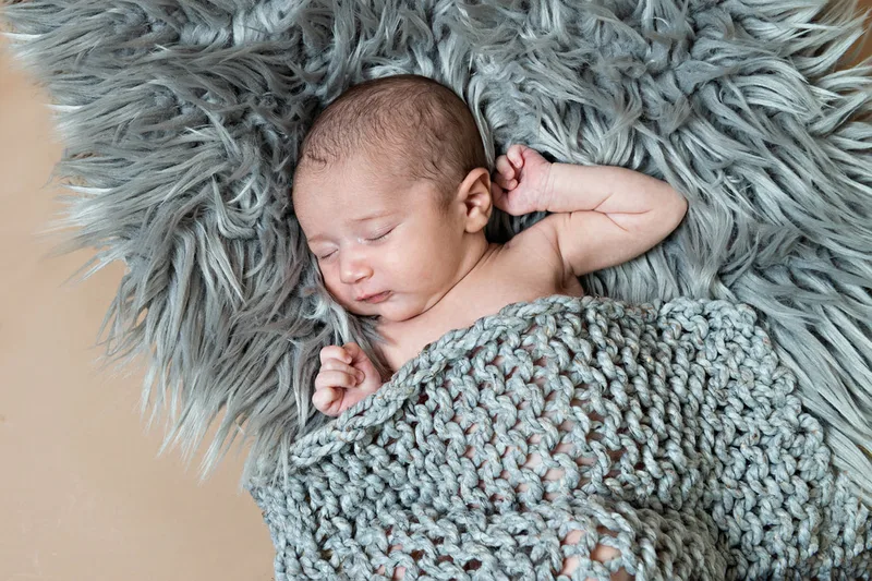 Color photo of a newborn baby boy covered in a gray knit blanket on top of a gray furry rug photographed in a studio. His eyes are closed. The photo was taken by Laura Pea Photography, a Long Island based newborn photographer.