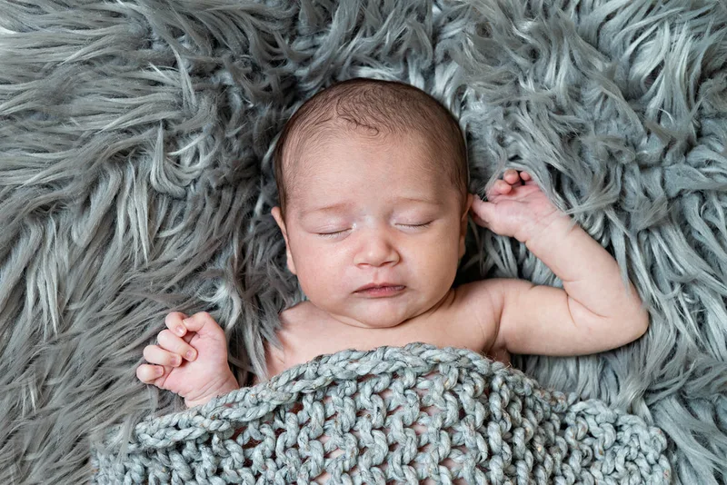 Color photo of a newborn baby boy covered in a gray knit blanket on top of a gray furry rug photographed in a studio. His eyes are closed. The photo was taken by Laura Pea Photography, a Long Island based newborn photographer.