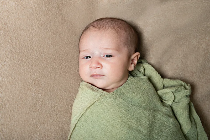 Color close up photo of a newborn baby wrapped in a green wrap on top of a beige blanket photographed in a studio. The photo was taken by Laura Pea Photography, a Long Island based newborn photographer.