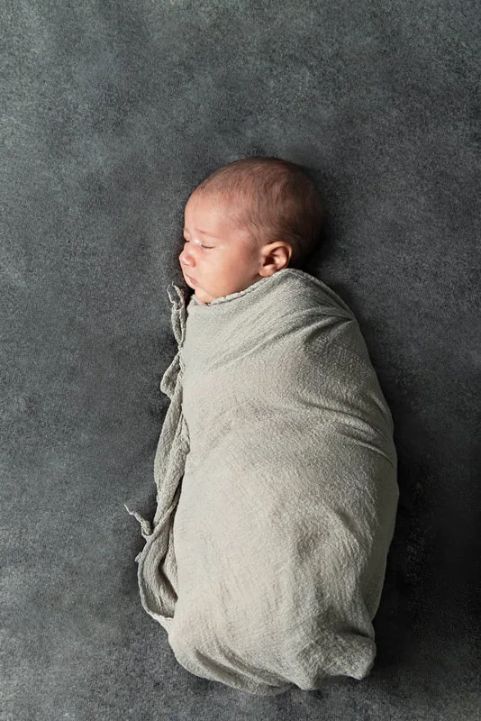 Color photo of a newborn baby boy wrapped in a gray wrap on top of a gray blanket photographed in a studio. His eyes are closed and he is laying in profile. The photo was taken by Laura Pea Photography, a Long Island based newborn photographer.