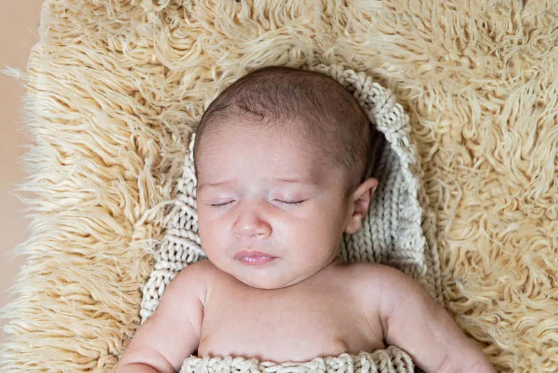 Color close up photo of a newborn baby boy covered in a beige cocoon wrap on top of a beige furry rug in a vintage doll bed photographed in a studio. His eyes are closed. The photo was taken by Laura Pea Photography, a Long Island based newborn photographer.
