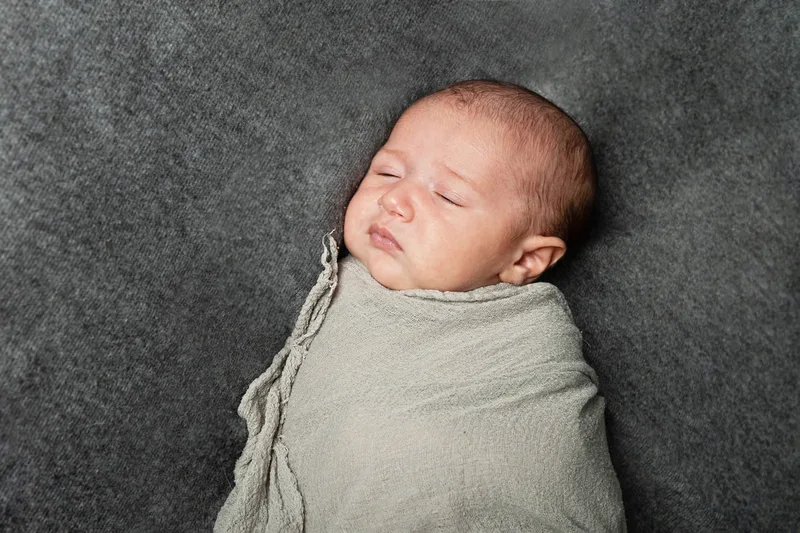 Color photo of a newborn baby boy wrapped in a gray wrap on top of a gray blanket photographed in a studio. His eyes are closed. The photo was taken by Laura Pea Photography, a Long Island based newborn photographer.