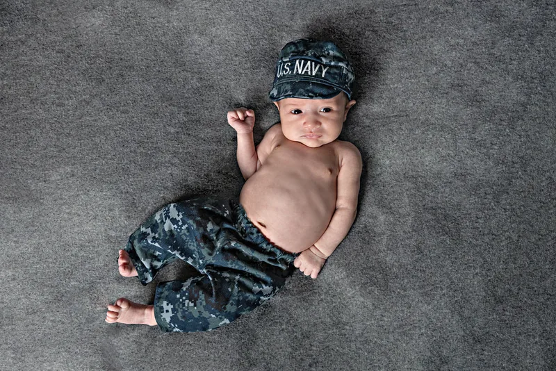 Color close up photo of a newborn baby dressed in a camouflage United State Navy outfit on top of a gray blanket photographed in a studio. The photo was taken by Laura Pea Photography, a Long Island based newborn photographer.