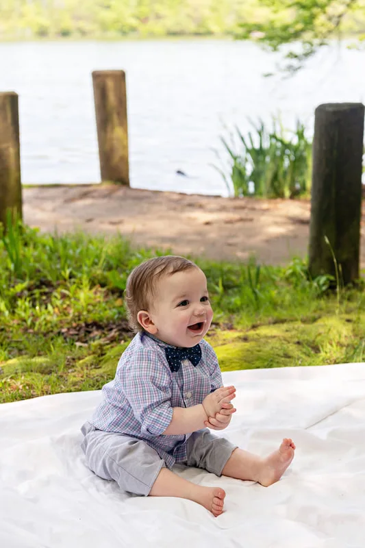 photo of a one year old baby boy dressed in a shirt and bow tie sitting on a blanket on a forest floor in front of a dock