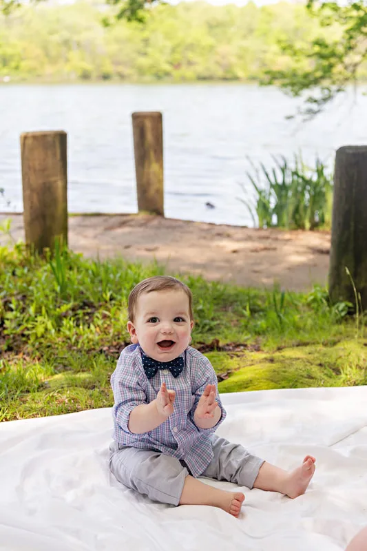 photo of a one year old baby boy dressed in a shirt and bow tie sitting on a blanket on a forest floor in front of a dock