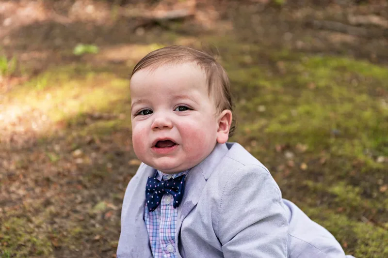 photo of a one year old baby boy dressed in a suit and bow tie sitting on a forest floor