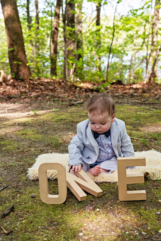photo of a one year old baby boy dressed in a suit and bow tie sitting on a blanket on a forest floor with brown craft letters that spell out the word "one"