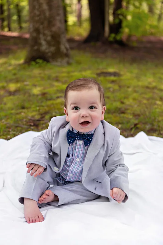 photo of a one year old baby boy dressed in a suit and bow tie sitting on a blanket on a forest floor