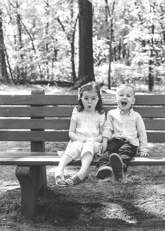 black and white photo of two four year old twin brother and sister sitting on a bench in a wooded park making silly faces
