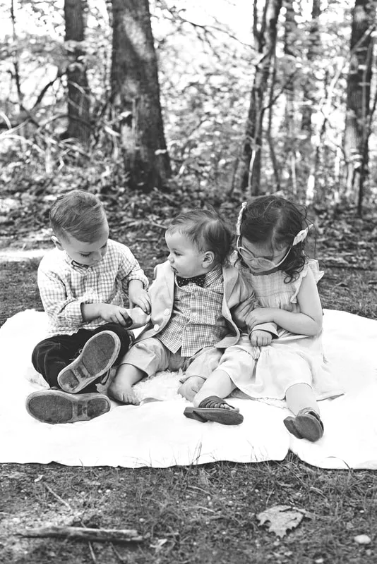 black and white photo of two four year old twin brother and sister with their one year old baby brother sitting on a blanket in a wooded park
