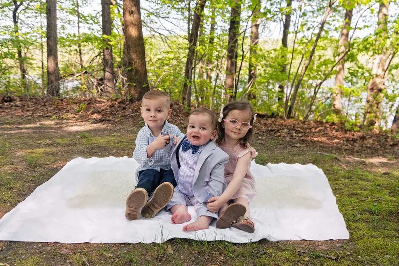 photo of two four year old twin brother and sister with their one year old baby brother sitting on a blanket in a wooded park
