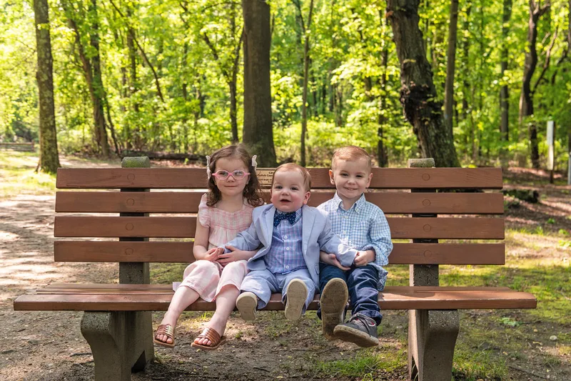 photo of two four year old twin brother and sister with their one year old baby brother sitting on a bench in a wooded park