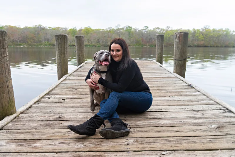 A color photo of a pit bull and her owner sitting together on a dock in front of a pond