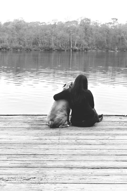 A black and white photo of a pit bull and her owner from behind sitting together at the end of a dock in front of a pond